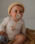A baby in a beige knit hat and cream onesie sits on a mustard blanket, holding a blue elephant teether and sucking on a FRIGG Daisy Silicone Pacifier (2-pack) by FRIGG, attached to a beaded clip made of medical grade silicone.