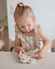 A young girl with light brown hair, wearing a striped dress with ruffled sleeves, sits at a table enjoying sensory play and building fine motor skills with the mushie Flower Press Toy, a pastel-colored flower-shaped fidget toy.