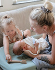 A woman with a bun and a forearm tattoo sits on a couch holding a newborn on the mushie Extra Soft Muslin Changing Pad Cover, while a young girl beside her reaches toward the baby with a small wooden toy.