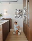 A toddler rides a mushie Mini Toddler Bike in a modern kitchen with wood and metal decor.