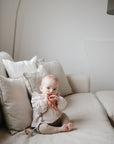 A baby sits on a beige sofa with cream cushions, holding and chewing on the mushie Daisy Rattle Teether in a softly lit, neutral-toned living room.