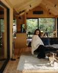 A woman with long hair, wrapped in a mushie Muslin Nursing Cover, sits on a sofa in a bright living room. A child's wooden bike rests on a white rug nearby, with trees visible through large windows.