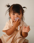 A young child in a beige shirt smiles at the camera indoors, holding a FRIGG Daisy Natural Rubber Pacifier from the FRIGG Daisy Natural Rubber Pacifier 2-Pack to their mouth.