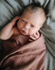 A newborn with light hair lies on a soft gray surface, wrapped in a mushie Organic Cotton Muslin Swaddle Blanket, gazing upward with a gently furrowed brow and one hand near their face.