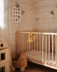 A cozy, sunlit nursery with a wooden crib, wicker pram, dollhouse, pastel bunting, and neutral decor. The mushie Star Lovey Blanket in organic cotton rests nearby as light streams in through a sheer white curtain.