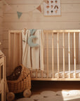 A cozy nursery features a wooden crib with the mushie Moon Lovey Blanket and a crescent moon pillow on the rail, beside a wicker toy stroller, with bunting and a wall poster adding charming decor.