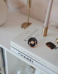 A close-up of The Kinfolk Table and Chanel Collections books stacked on a white shelf features the FRIGG Daisy Natural Rubber Pacifier by FRIGG resting on top, complemented by a gold lamp and a white vase for a stylish touch.
