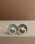 The FRIGG Daisy Night Natural Rubber Pacifier 2-Pack by FRIGG, in light blue and taupe, features scalloped edges, a minimal modern design, and natural rubber latex nipples. Shown resting on a beige surface with a soft brown background.
