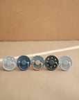 Five FRIGG Moon Natural Rubber Pacifier 2-Packs by FRIGG, in pastel shades, stand upright on a beige surface with soft lighting, showcasing a minimalistic baby essentials aesthetic.