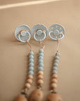 Three blue pacifiers are attached to mushie’s Silicone Pacifier Clip | Eva, featuring food-grade silicone and natural wood-colored beads, all shown on a beige background.