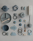 A neatly arranged set of blue and gray mushie baby items—plates, spoons, snack cups, a Finger Toothbrush, pacifiers, teethers, a beaded pacifier clip, and packaging—displayed on a light background.