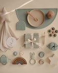 A flat lay of pastel-colored baby items on a beige background, featuring the mushie Flower Press Toy along with a star plush, silicone plates, rainbow stacker, utensils, teether, bib, pacifier, rattle, and soft cloth.
