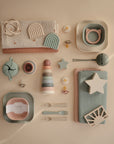 A flat lay of pastel-colored baby items, including non-toxic plates, forks, cups, pacifiers, folded cloths, teething toys, and the mushie Stacking Rings Toy, all neatly arranged on a beige background.