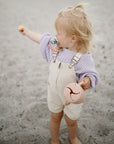 A young blond child in a lilac sweater and beige overalls stands barefoot on sand, holding a pink snack cup with the mushie Silicone Pacifier Clip | Eva and pointing to the side.
