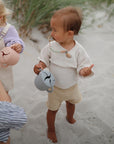 Two young children stand barefoot on sandy grass, holding pastel cups with bird designs. One child, wearing the mushie Silicone Pacifier Clip | Eva, looks to the side while an adult’s hand is partially visible.