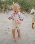 Two young children stand barefoot on a sandy beach, each holding a cup secured with the mushie Silicone Pacifier Clip | Eva—practical pacifier accessories for carefree play. Their lavender and beige outfits add to their charming look. Grass waves in the background.