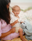 A smiling toddler sits on an adult's lap at the beach, holding a mushie Snack Cup. The adult wears a pink floral dress, and both are on a yellow blanket with sand and grass in the background.