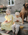 Two young children sit on wooden steps outside with a large brown dog. Nearby, mushie Round Dinnerware Bowls made from BPA-free plastic rest on the steps near the kids.