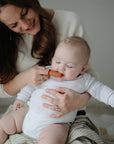 A smiling woman helps a baby in white use the mushie Finger Toothbrush, a brown silicone brush, making baby teeth cleaning easy and enjoyable. Both look relaxed and content in a softly lit room.