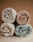 Four rolled muslin blankets in cream, beige, light blue, and stripes are stacked together against a plain brown background, resembling the mushie Extra Soft Muslin Changing Pad Cover.