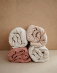 Four rolled towels in beige and dusty pink are neatly stacked, echoing the soothing palette of the mushie Extra Soft Muslin Changing Pad Cover, set against a plain light brown background.