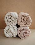 Four rolled-up swaddles in neutral tones sit in a square—three solid beige and brown, one white with brown florals—beautifully matching your mushie Extra Soft Muslin Changing Pad Cover against a beige background.