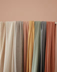 The mushie Baby Wrap in soft, muted colors such as beige, blush, and rust is draped over a white rod against a pale pink background.
