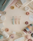 A flat lay of pastel mushie baby products, including the Flower Training Toothbrush, creams, stacking toys, teethers, skincare bottles, and soft cloths, all neatly arranged on a beige surface.