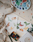 A flat lay featuring watercolor dinosaur art, paint supplies, and baby essentials—including a mushie Organic Cotton Muslin Swaddle Blanket with dinosaur prints—on a beige surface decorated with small colorful dinosaur illustrations.