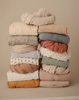 A stack of mushie Mini Muslin Crib Sheets, crafted from 100% cotton muslin in soft, earthy tones and patterns, neatly folded against a plain beige background.