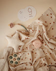 A soft, patterned muslin blanket is laid out with baby toys, a star cushion, a mushie Silicone Pacifier Clip (Eva), a pop-it toy, and milestone cards—including one marked 9 months—displayed on a beige background.