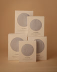 Five cream-colored mushie Extra Soft Bassinet Muslin Crib Sheet boxes, each with a circular window showing light gray fabric, are stacked against a beige background—perfect for breathable baby bedding.