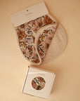 A partly folded floral-patterned mushie Organic Cotton Muslin Burp Cloth from the 2-Pack set rests on a beige background, with its matching box below. Both feature the mushie label and are crafted from soft organic cotton.