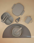 A set of gray silicone baby feeding items—bib, divided plate, bowls, easy-grip cutlery, Mushie Trainer Sippy Cup (non-toxic), snack container, and half-circle placemat—are neatly arranged on a light beige surface.