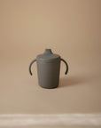 A gray Mushie Trainer Sippy Cup with easy-grip handles and a spout, featuring the embossed "mushie" logo, is centered on a beige surface against a soft, blurred background.