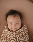 A baby with curly hair smiles softly while lying on a light brown surface, wrapped in the mushie Extra Soft Bassinet Muslin Crib Sheet in a leopard-print design.