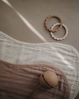Two scalloped-edge teething rings and a beige pacifier rest on mushie's Organic Cotton Muslin Burp Cloth 2-Pack in neutral tones, displayed on a light brown surface under soft natural light.
