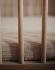 Close-up of mushie’s Extra Soft Bassinet Muslin Crib Sheet in beige, visible through natural wood crib bars and showcasing its breathable texture in a minimal, neutral-toned nursery setting.