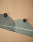 Two overlapping mushie Silicone Placemats in muted green, made from food-grade silicone, are secured to a tan surface with black round magnets. Each has an embossed “mushie” logo, and a diagonal stripe of light runs across both.