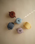 Five mushie Snack Cups in brown, blue, yellow, gray, and lavender sit on a beige surface. Each silicone toddler cup has handles and a spill-proof slotted lid. Soft light with a sunbeam illuminates the scene.
