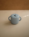 The mushie Snack Cup is a small, light blue silicone cup with two handles and a flexible petal-like opening—designed to be spill-proof and perfect for toddlers. It sits on a beige surface against a brown background.