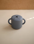 The mushie Snack Cup is a small, light gray silicone cup with two handles and a soft, petal-shaped spill-proof lid, shown on a cream surface against a brown background—ideal for toddlers.