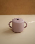 The mushie Snack Cup is a small, light purple silicone cup with two handles and a flexible, petal-like spill-proof lid—ideal for toddlers. It’s shown on a beige surface with a blurred brown background.
