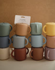 Twelve Snack Cups by mushie, in vibrant silicone colors and with handles, are stacked in three columns against a brown background. A card labeled “mushie” is placed between two top cups, showcasing these toddler snack containers.