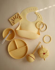 A pale yellow mushie Snack Cup for toddlers is displayed on a neutral background alongside matching silicone feeding items like a bib, divided plate, spoon, teether rings, rattle, and cups.