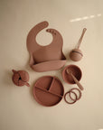 The mushie Snack Cup in dusty rose, shown with matching silicone bib, spoon, bowl, divided plate, teether, and beaded accessory—perfect for toddlers and neatly arranged on a light beige background.