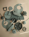 A flat lay of assorted mushie silicone baby feeding items in muted green, featuring the spill-proof Snack Cup for toddlers, along with bibs, divided plates, spoons, bowls, a cup, teether, brush, and snack containers on a beige surface.