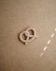The mushie Pretzel Teether, a small light beige silicone teether shaped like a pretzel, rests on a plain brown textured surface with diagonal sunlight casting spots of light on the right side.