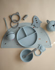 The mushie Snack Cup and other light blue silicone baby feeding items—sectioned plate, bowl with spoon, bib, handled cup, placemat, and teether—are neatly displayed on a beige surface.
