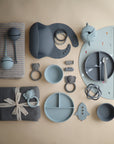 A neatly arranged flat lay of mushie baby and toddler feeding items in blue and gray, featuring plates, bowls, spoons, a silicone Snack Cup, bib, teether rings, muslin cloths, and spill-proof accessories on a beige background.